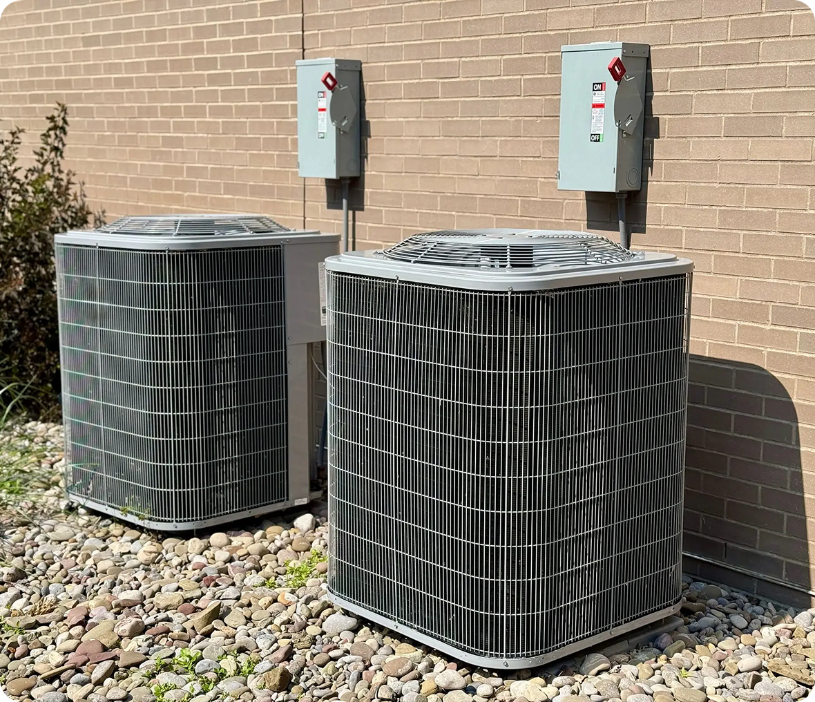 Two air conditioners outside of a brick building, surrounded by stones and plants