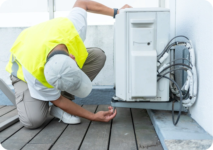 Man in a yellow vest and hat working on an air conditioner on a wooden surface