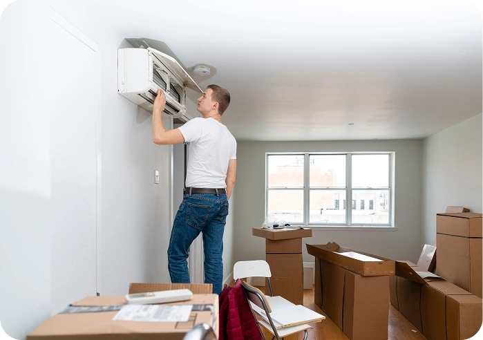 Man standing on the floor, holding an air conditioner in his hand, surrounded by cardboard boxes, chairs, and other objects